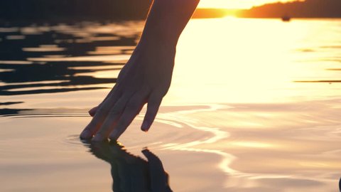 Close-up of a woman hand gently touches the surface of the water in the sea or lake, slowly leads her hand on the water and raises up, in the warm golden rays of the sun at sunset in summer evening.