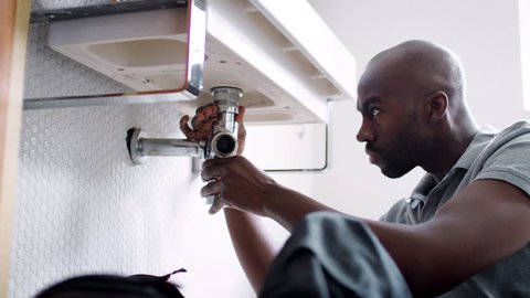 Young black male plumber sitting on the floor replacing the trap pipe under a bathroom sink, low angle