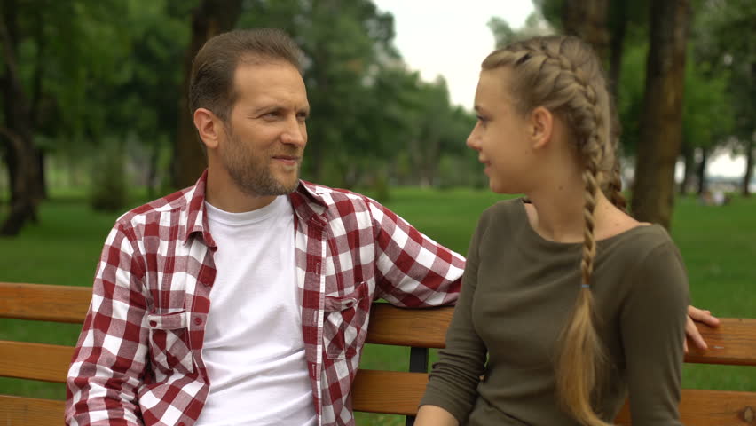 Beautiful teenage daughter whispering secrets to father, relax on bench in park