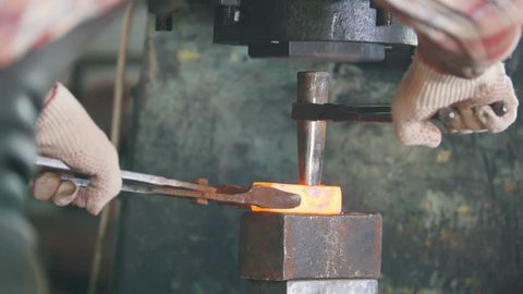 Blacksmith at work with electric hammer on the anvil, making hole in red hot steel, craft