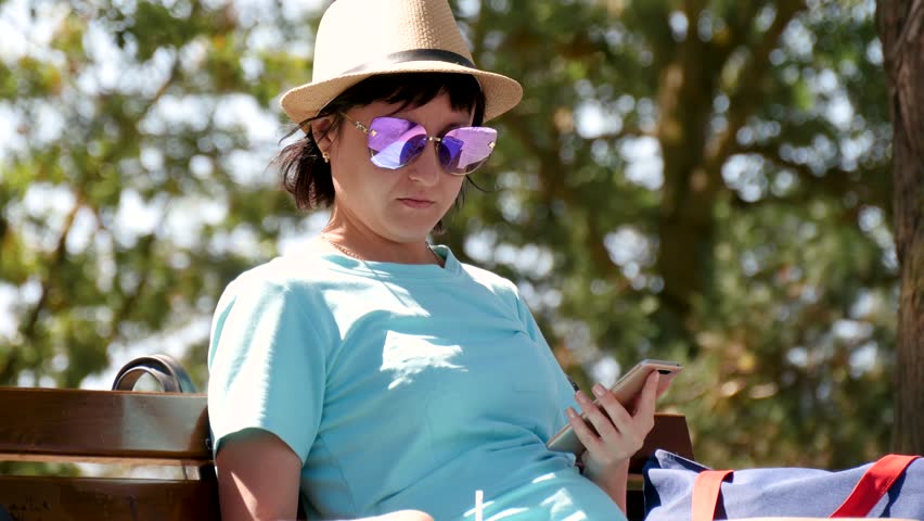 Brunette in a hat and dress sits on a park bench, drinks coffee and uses the phone.