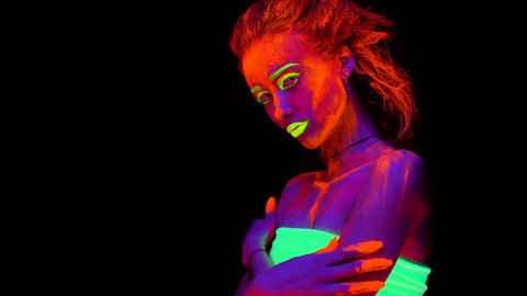 A beautiful young sexy girl with ultraviolet paint on her body looking looking into camera and turning from side to side. Pretty woman with glowing bodyart in black lamp light.