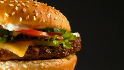 Juicy beef burger with cutlet, onion, vegetables, melted cheese, lettuce, sauce and topped sesame seeds. Isolated hamburger rotates on dark background, close-up view