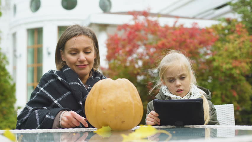 Mother carving pumpkin while daughter looking for tips in internet, blogging