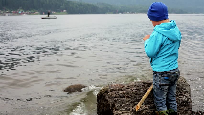 summer landscape on the lake in the mountains. one little boy in a blue jacket and rubber boots playing on the shore. the stick hits the water