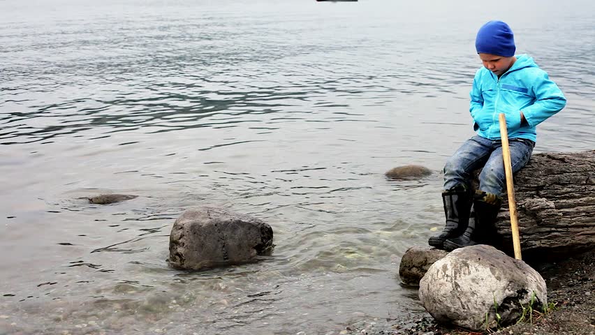 summer landscape on the lake in the mountains. one little boy in a blue jacket and rubber boots playing on the shore. the stick hits the water