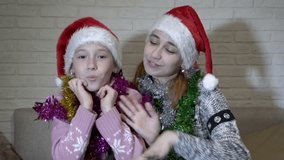 Portrait of two happy cheerful children who sit in Santa's hats with Christmas tinsel on their shoulders. They dance, laugh, move to the music and look at the camera. Close up. 4K. 25 fps