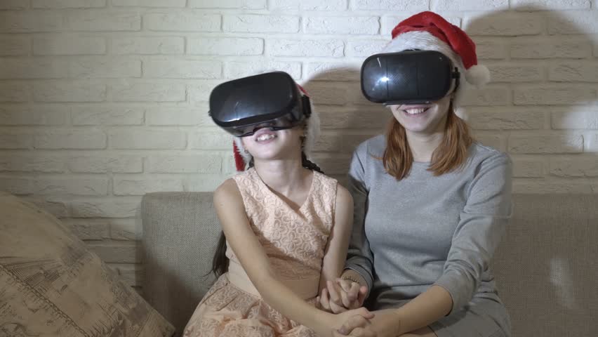 Happy kids in virtual reality helmets and Santa Claus hats on their heads sit and play at home on the couch against a white brick wall. Close up. 4K. 25 fps