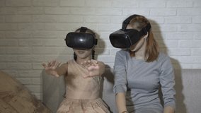 Children play virtual games. Two sisters in virtual reality glasses sit and play at home on the couch against a white brick wall. They wave their hands and look around. Close up. 4K. 25 fps