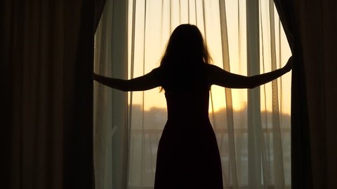 Young woman open curtains and look to beautiful prime view, half hour before sunrise, bright sky and hill silhouette seen blurred outside. Dark girl silhouette against transparent hangings
