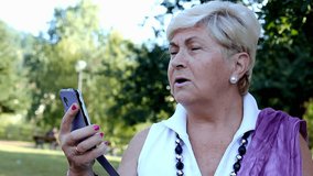 Blond senior lady talking on cellphone with loud speaker. Elder woman with sleeveless white shirt holding smart phone in the park. Conversation, communication concepts - Powered by Shutterstock - Get 15% off with code: PIKWIZARD15