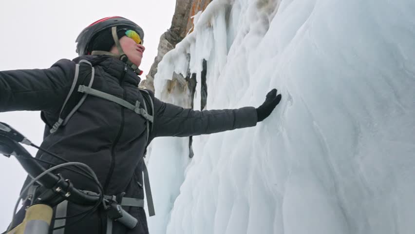 Man is walking beside bicycle near the ice grotto. The rock with ice caves and icicles is very beautiful. The cyclist is dressed in gray down jacket, cycling backpack and helmet. The tires on covered