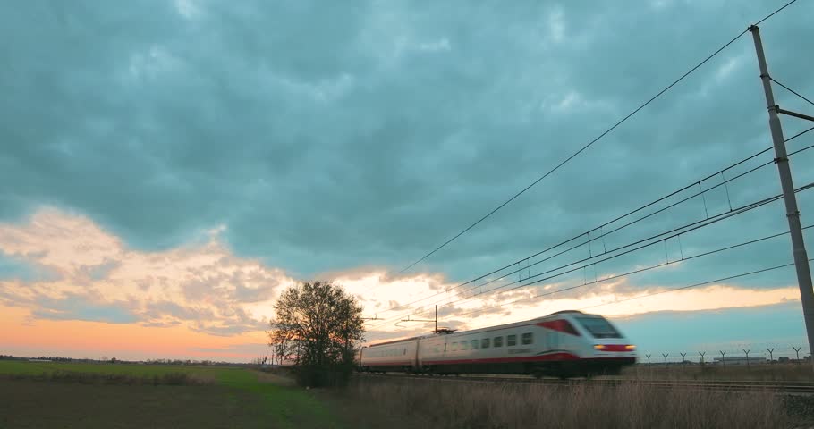 Passenger white and red high speed train is passing under a cloudy sky at sunset