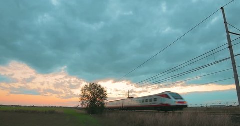 Passenger white and red high speed train is passing under a cloudy sky at sunset