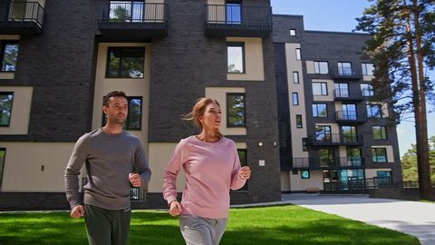 Girlfriend and boyfriend jogging outdoor slow motion near with modern apartment block. Freshness sport concept of spring recreation and body caring. Active beautiful young couple running in park
