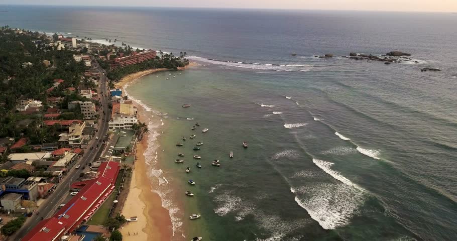 Magnificent sunny landscape of the sand seashore of Hikkaduwa on the sky background on Sri Lanka. There are many boats swinging on waves, buildings and roadways with traffic between green trees.