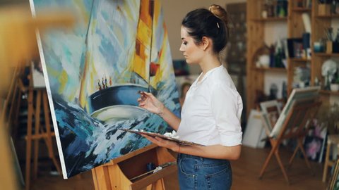 Female artist is concentrated on work painting beautiful picture sea and boat with oil paints working alone in studio using bright colors. Artistry and creativity concept.
