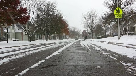 Establishing shot of a street in typical suburban American neighborhood during snow fall in winter, with houses and trees on both side and lawns covered with snow. A school symbol is also visible