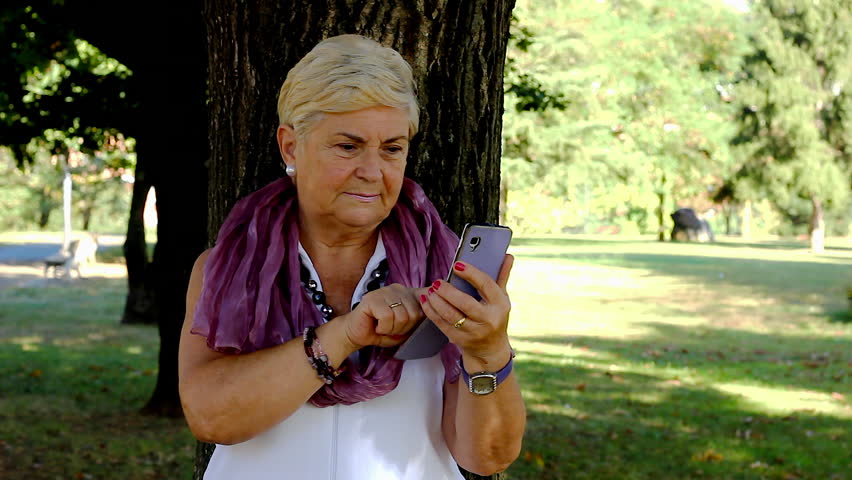 Euphoric senior woman celebrating sport victory looking at smart phone at park. Lucky mature lady acting exultant holding mobile on sunny day outdoors. On line bet win concept Royalty-Free Stock Footage #1019618902