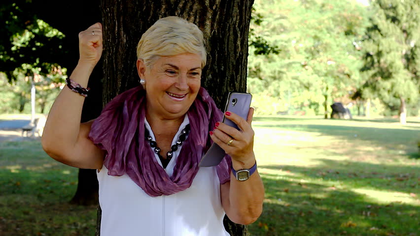 Euphoric senior woman celebrating sport victory looking at smart phone at park. Lucky mature lady acting exultant holding mobile on sunny day outdoors. On line bet win concept Royalty-Free Stock Footage #1019618902