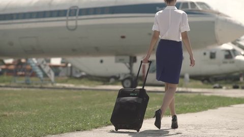 Beautiful and cheerful flight attendant walks towards the airplane. Stewardess with a suicase. Highheal shoes. Sexy flight attendant.