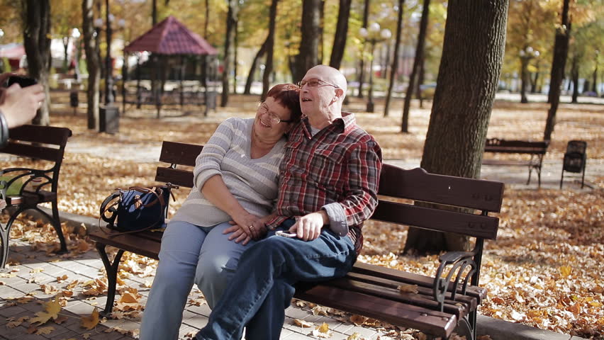 Adult son takes pictures of elderly parents on smartphone sitting on bench in autumn Park.