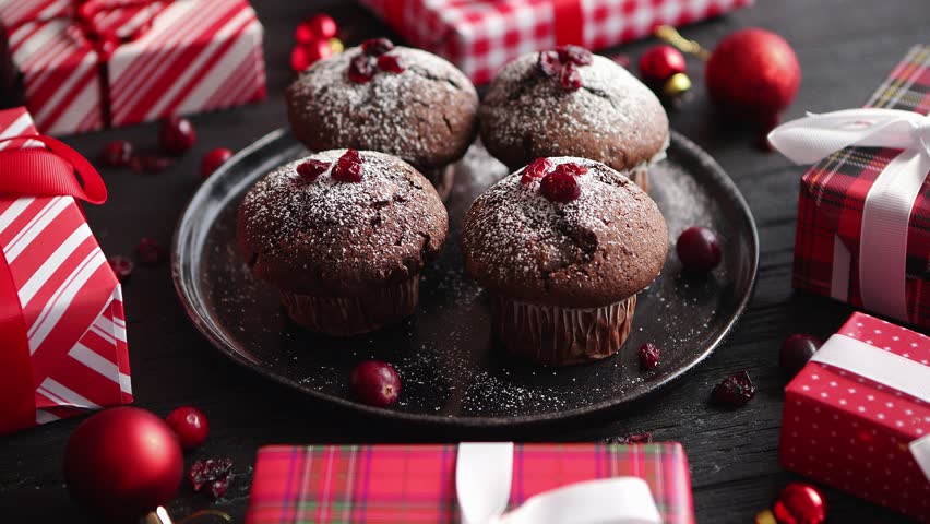 Christmas chocolate delicious muffins served on black ceramic plate. Sprinkled with powder sugar. Cranberries on top. Xmas gifts and decorations on sides.