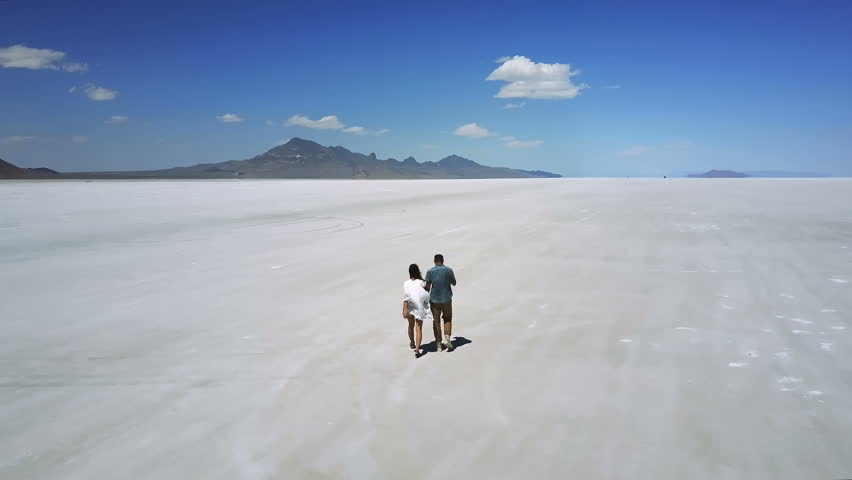 Drone follows romantic couple holding hands, walking towards epic white flat land in the middle of Utah salt desert lake