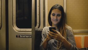 Happy relaxed beautiful millennial girl sitting in the metro train watching videos online on smartphone app and smiling.