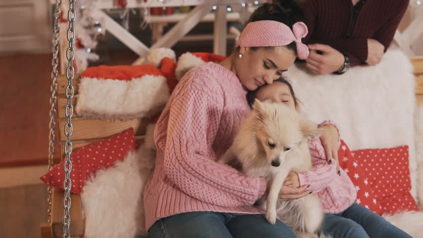 Young family with little daughter and white dog have fun sitting at Christmas bench. Happy family in pullovers celebrating winter holiday at porch, expressing love and care in slow motion. Medium shot