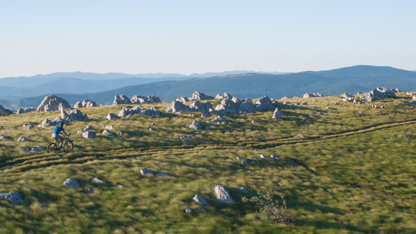 AERIAL: Athletic young man enjoying the pleasant summer weather by riding his bicycle along a scenic trail overlooking the vast green valley in Slovenia. Extreme mountain biker speeding down a hill.