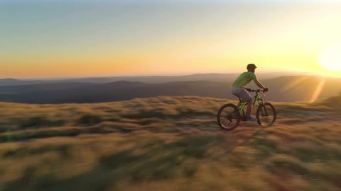 DRONE, LENS FLARE: Athletic male tourist speeding downhill on his cool mountain bike at sunset. Golden morning sunbeams illuminate the picturesque mountains and mountain biker riding down a trail.