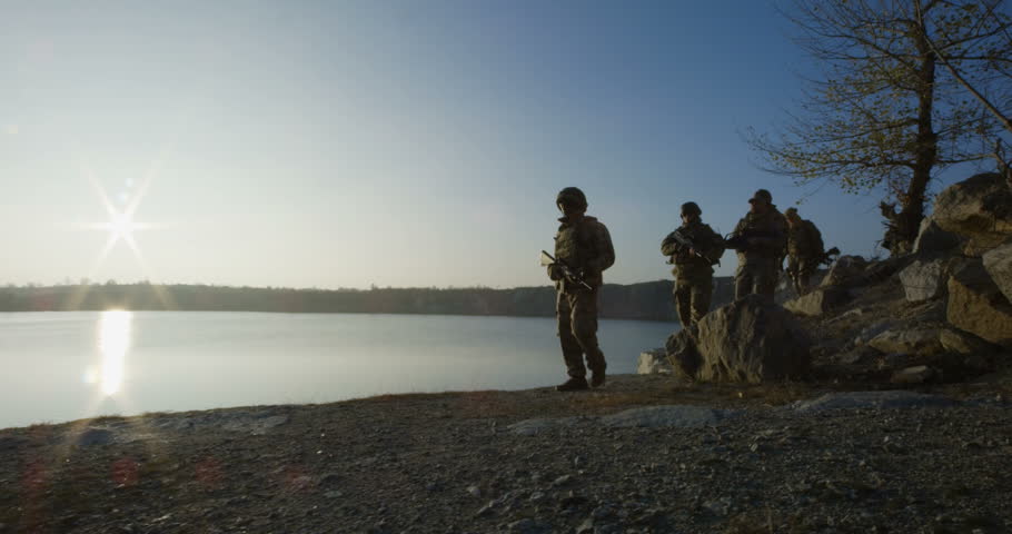 Medium slow motion shot of fully equipped and armed soldiers walking in single file by the side of a lake at sunset | Shutterstock HD Video #1019663062