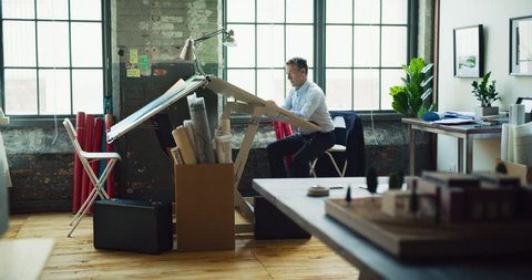 Architect man sits at drafting table in modern industrial office during the day. Wide to long shot on 4K RED camera on a gimbal.