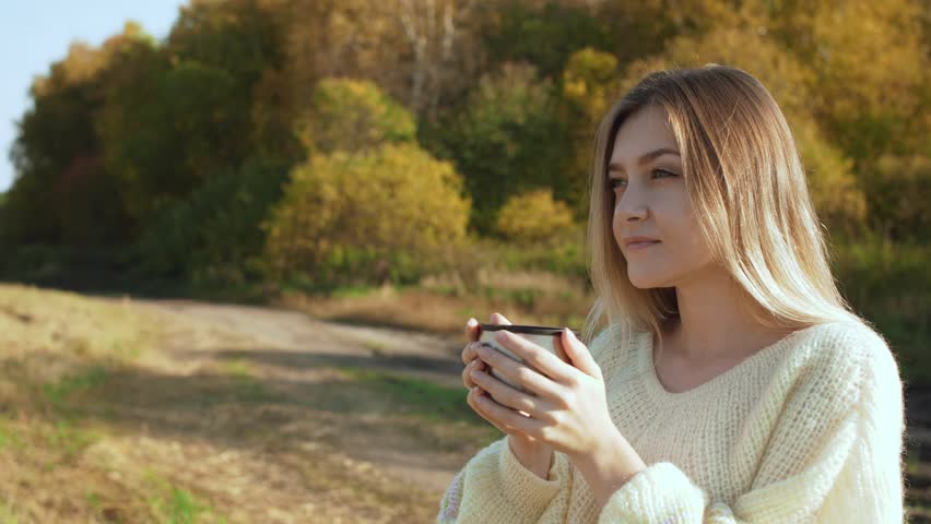 Young beautiful woman drinking hot tea from thermos cup. Over autumn nature background