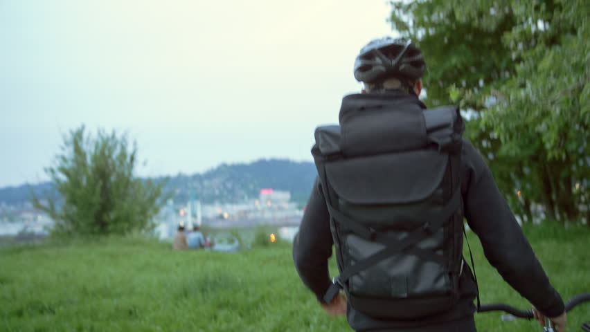Handheld rear view of male commuter with bicycle walking on grassy field against sky