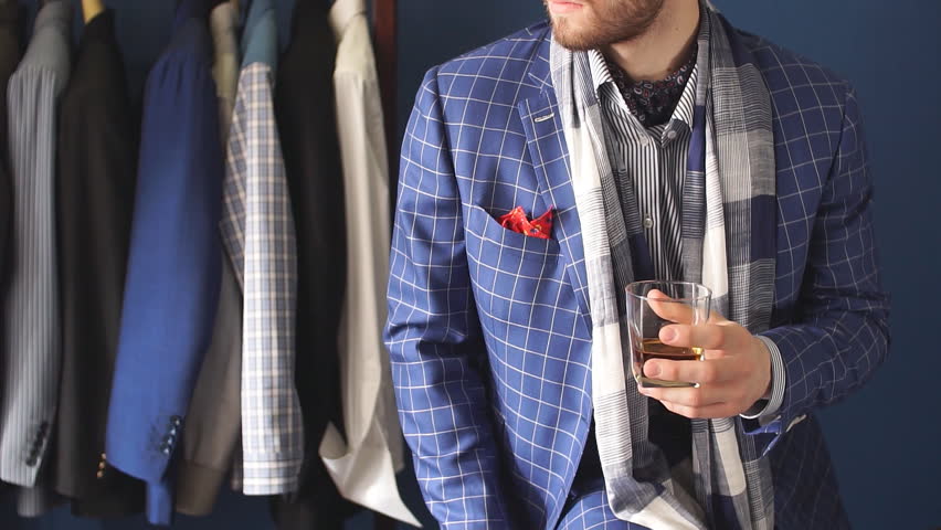 Elegant man in fashion checkered suit and hat posing at atelier with beverage in hand , clothes rack with ready made mans suits on background