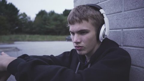 Close-up of a male teenager suffering from depression. He is sitting against a brick wall while listening to music through a pair of headphones.