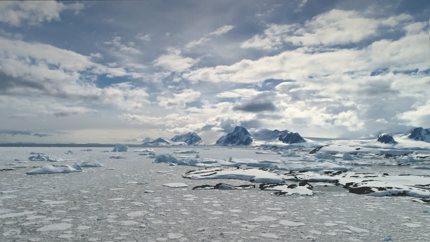 Aerial Flight Over Antarctica Ocean. Time Lapse. Hyper Lapse. Fast Moving Clouds On The Sky. Drone Overview Of Icebergs, Ice Pieces Floating In Sunlit Ocean. Snow Covered Mighty Mountains Background