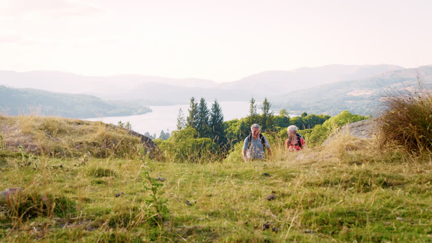 Slow Motion Shot Of Senior Couple Climbing Hill On Hike Through Countryside In Lake District UK Together