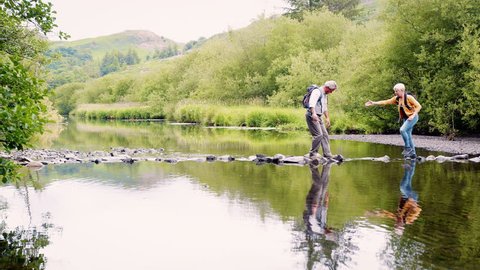 Slow Motion Shot Of Senior Couple Using Stepping Stones To Cross River Whilst Hiking In UK Lake District