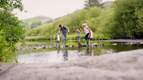 Slow Motion Shot Of Parents Helping Children To Cross River Whilst Hiking In UK Lake District
