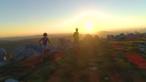 DRONE, SUN FLARE: Flying behind sporty tourist couple running along mountain trail at sunset. Golden morning sunbeams shine on athletic young man and woman training for a difficult fell running race.