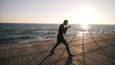 Full lenght of a young athlete in black shirt and red sneakers while training process on the promenade in front the sea in the early morning. Man boxing with invisible opponent, punching wearing black
