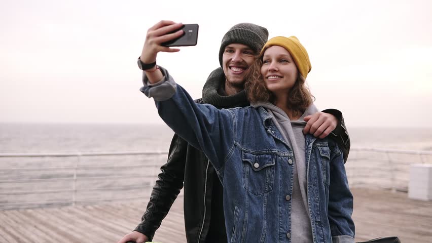 Happy young couple taking selfie by mobile phone near the seaside in cloudy day. Girl in yellow hat holding the smartphone, making funny faces. Beautiful couple selfie, love and bicycle concept