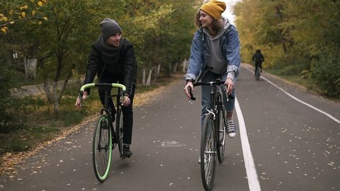 Young, smiling friends or young couple in hats cycling on their trekking bikes through the autumn park on bikes. Man and woman riding bikes together and talking. Front view