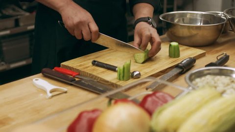 Professional chef cutting slices of cucumber on a cutting board in industrial kitchen with soft interior lighting. Close up shot on 4k RED camera.