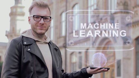 Smart young man with glasses shows a conceptual hologram Machine Learning. Student in casual clothes with future technology mobile screen on university background