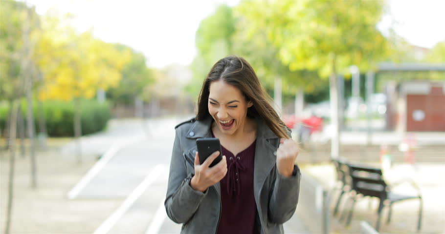 Front view of an excited woman walking towards camera, checking phone and celebrating good news in a park