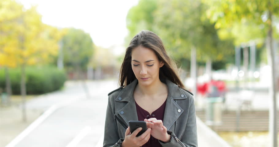 Front view of a perplexed woman walking checking smart phone content and then stops looking at camera in a park | Shutterstock HD Video #1019750350
